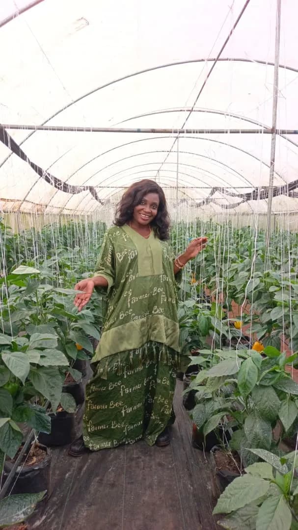 A smiling member standing between rows of pepper plants inside her greenhouse, one hand held out toward the plants.